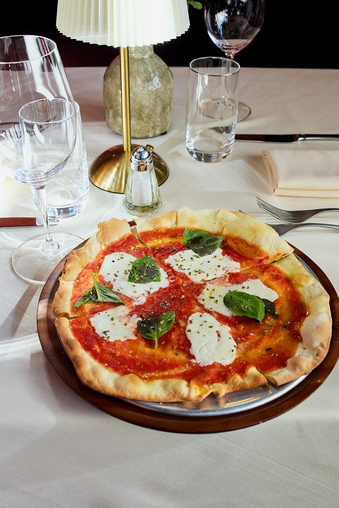 A thin-crust pizza with vibrant tomato sauce, fresh mozzarella rounds, and basil leaves. It sits on a white tablecloth next to wine glasses and a gold table lamp.