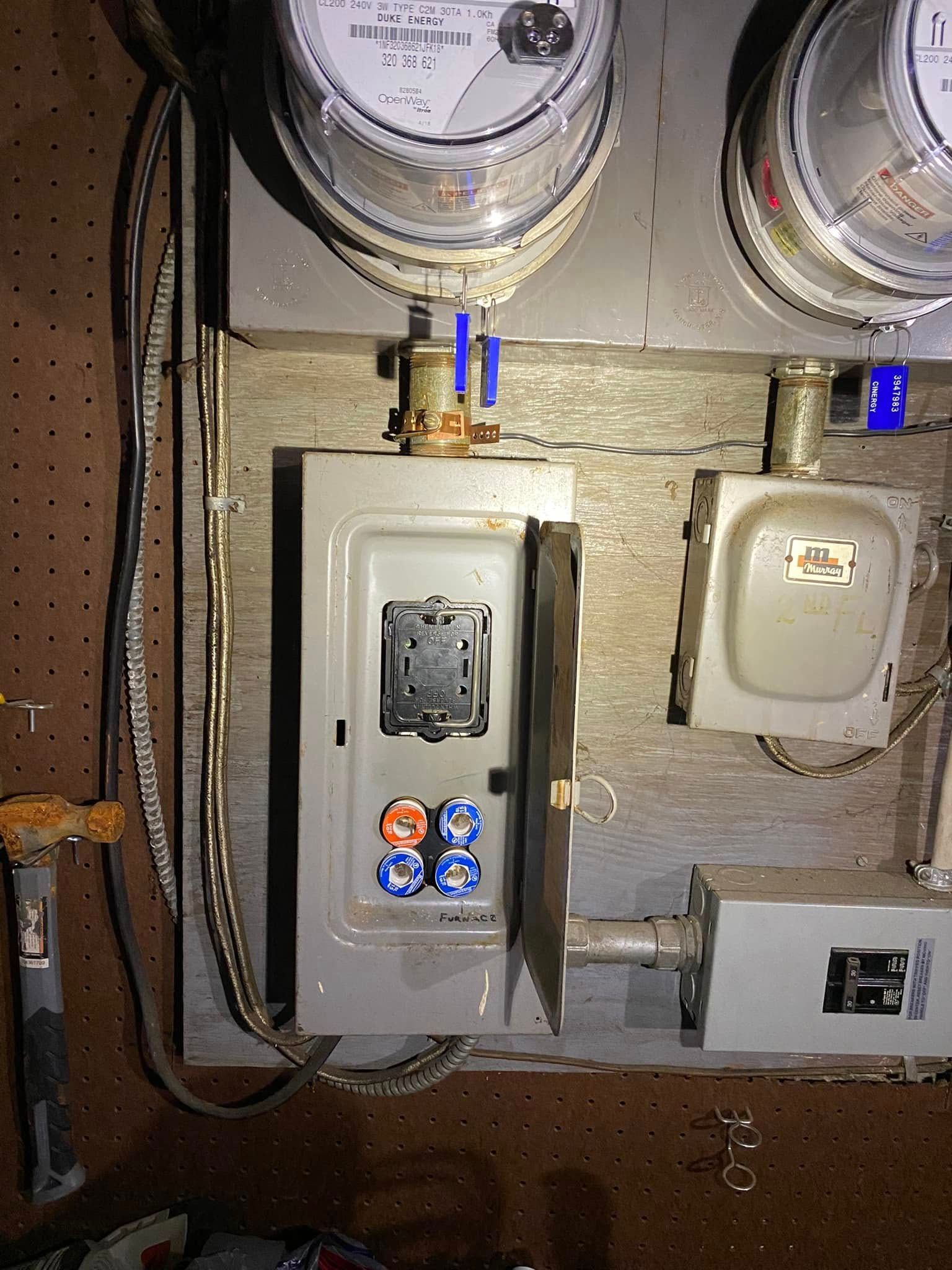 Electrical meter and fuse panel mounted on wall with visible wiring, conduits, and service boxes, showing older installation setup with labeled components, exposed cables, and utility connections in basement environment.