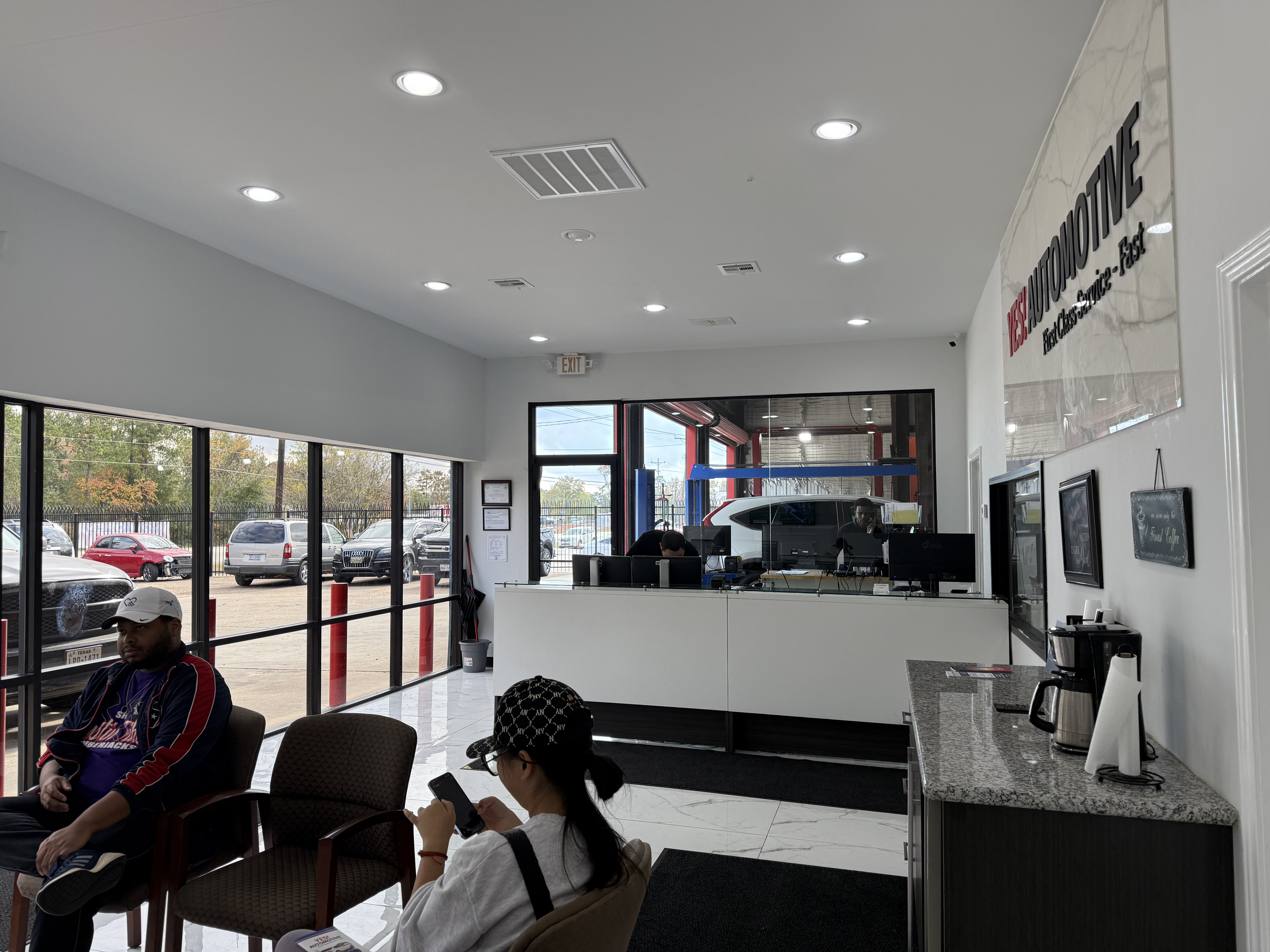 Interior view of YES! Automotive Porter, TX customer waiting area and service reception. The facility features a bright, modern lobby with comfortable seating, a spacious service counter, large windows, complimentary coffee, and a clear view of the automotive repair bays. Customers can relax while service advisors assist with vehicle maintenance and repair needs.