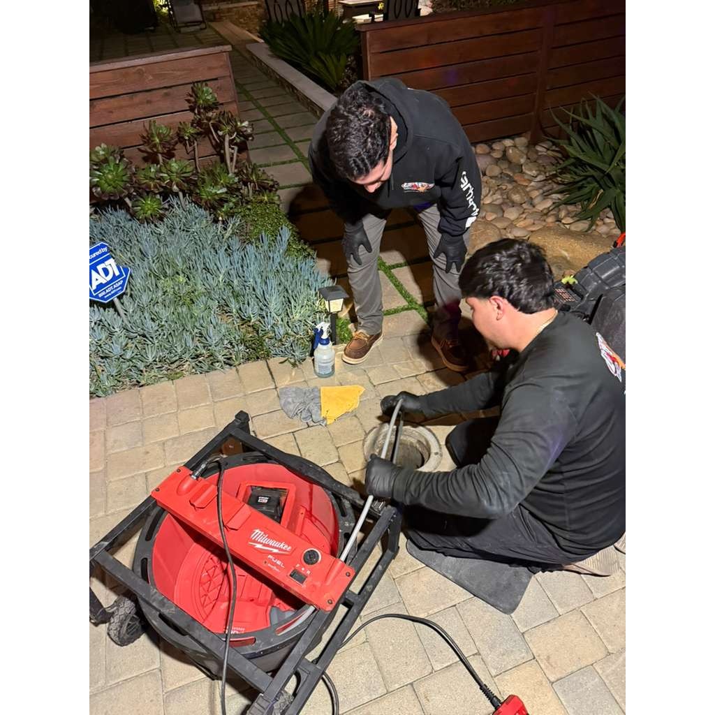 Two Pop & Go Rooter technicians work together to clear a drain at night using a powerful drain cleaning machine. The teamwork and specialized equipment highlight the company's commitment to solving plumbing problems efficiently, no matter the time of day.