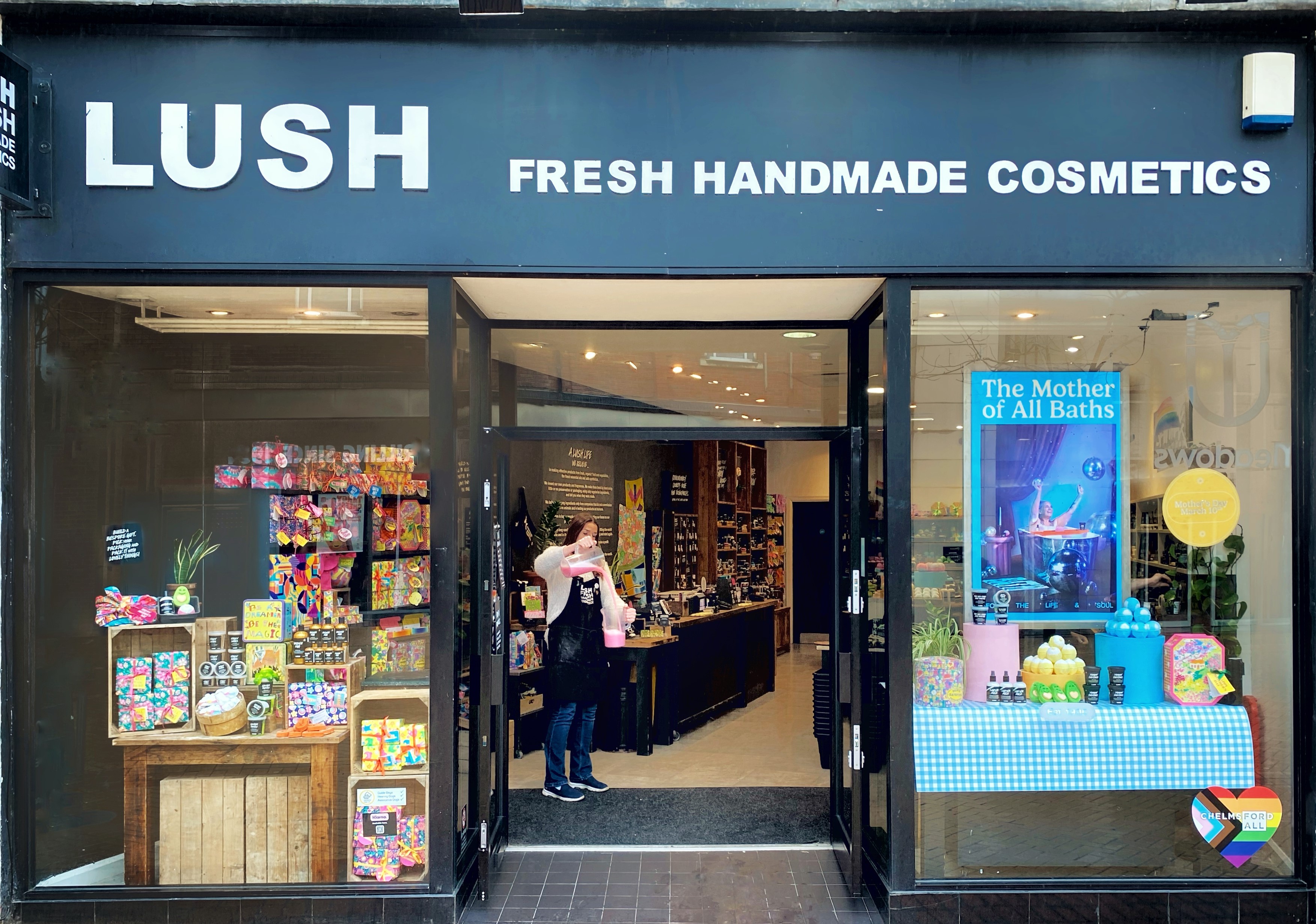 An image of Lush Chelmsford  from the outside of the store.  At the top of the image, "Lush Fresh Handmade cosmetics" is written in white text against a black background. On the left and right of the image, colourful Lush products are displayed in the window. A person is demoing bubble bars in bubble jugs at the front of the shop.