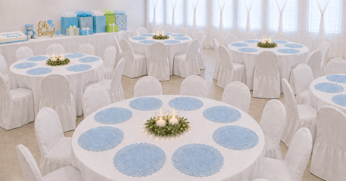 A beautifully decorated room for a baby shower, with round tables draped in white tablecloths and chairs covered in white slipcovers. Each table is set with pale blue placemats and a centerpiece of two lit candles surrounded by greenery and fairy lights. A table at the back is adorned with baby shower gifts in blue and green packaging, a teddy bear, and a 'Oh Baby!' sign.