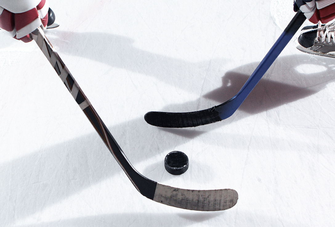 Close-up of two hockey sticks and a puck on an ice rink. The sticks, held by players in skates, hover above the ice, ready for a face-off.