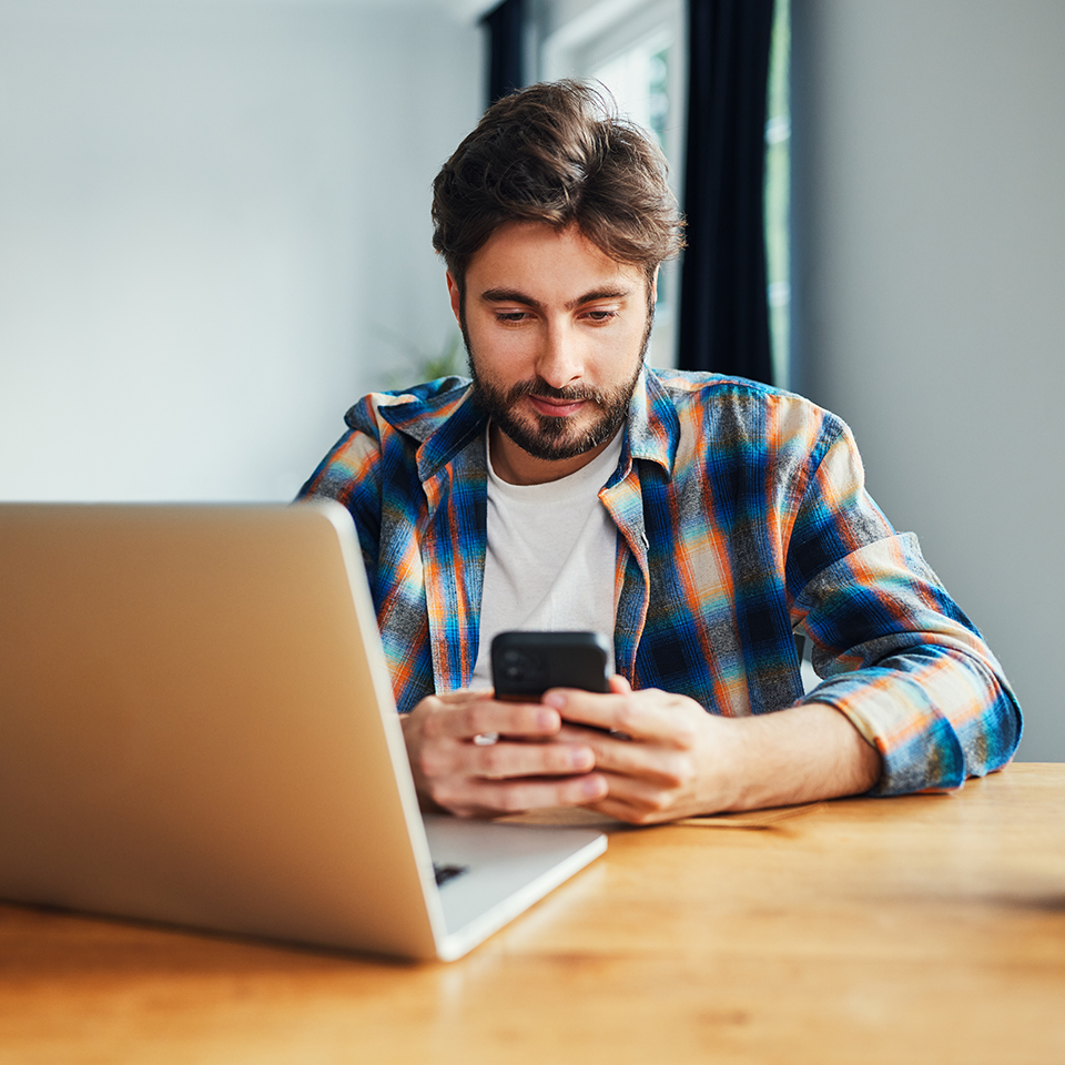 A man looking at a phone with a laptop open in front of him