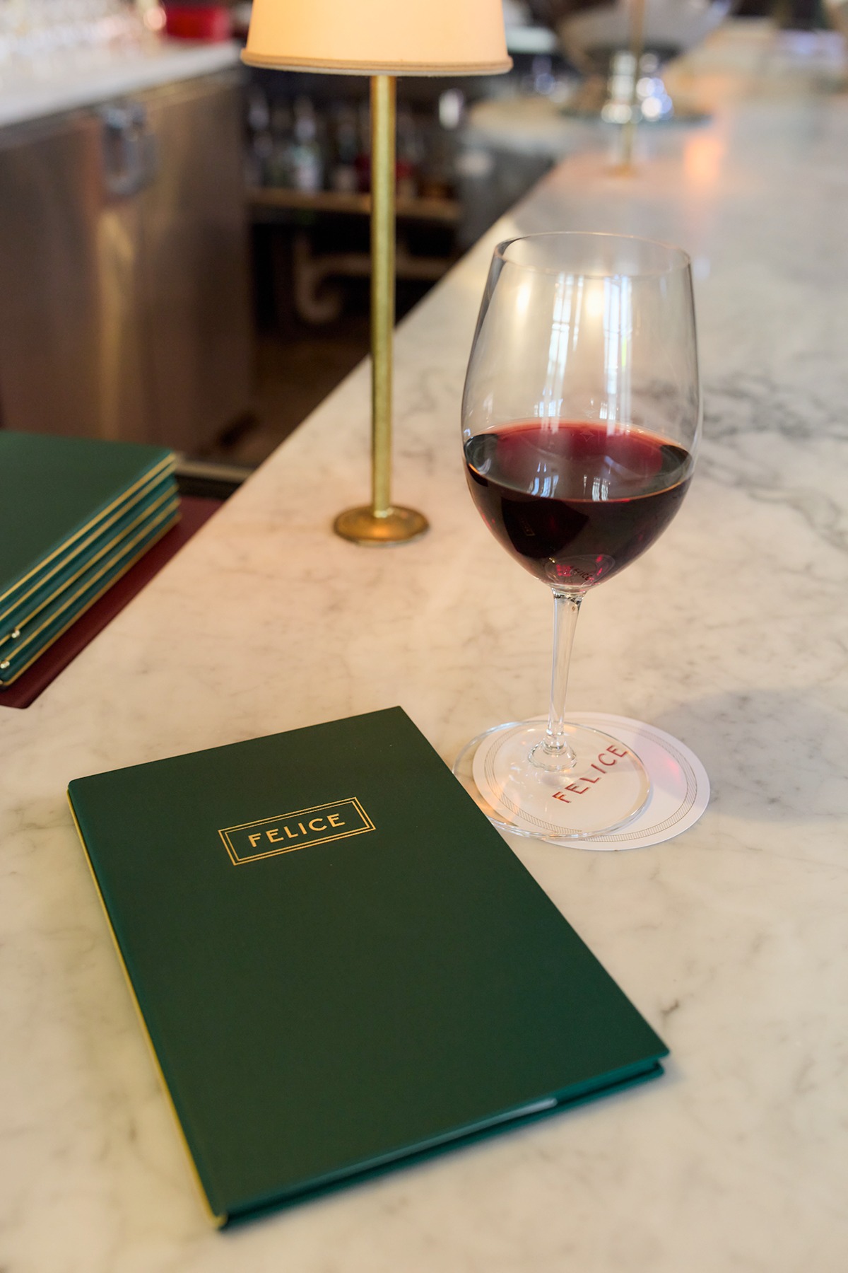 A glass of red wine sits on a Felice-branded coaster next to a dark green leather menu with gold lettering. The setting is a bright marble bar top with a vintage-style brass lamp in the background.
